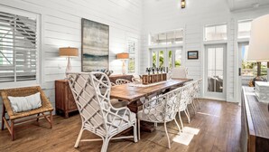 A spacious dining area with a wooden table and white wicker chairs, white paneled walls, and large windows. - Golden Daze 239 SE (Port Aransas)