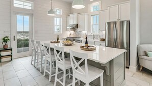 Une cuisine moderne avec des meubles blancs, un grand îlot central doté de tabourets, des appareils en acier inoxydable et un éclairage suspendu.