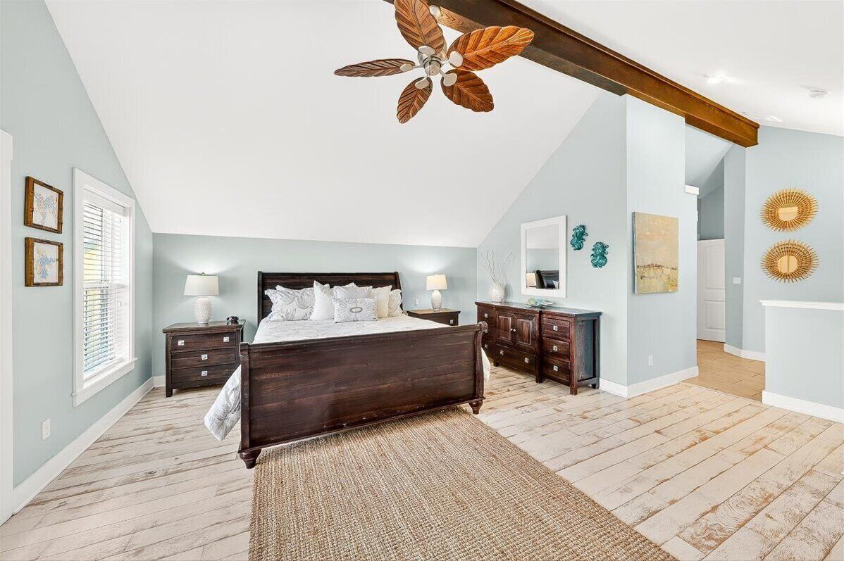 Quarto com cama de madeira, ventilador de teto, mesa de cabeceira, cômoda e uma grande janela com persianas.