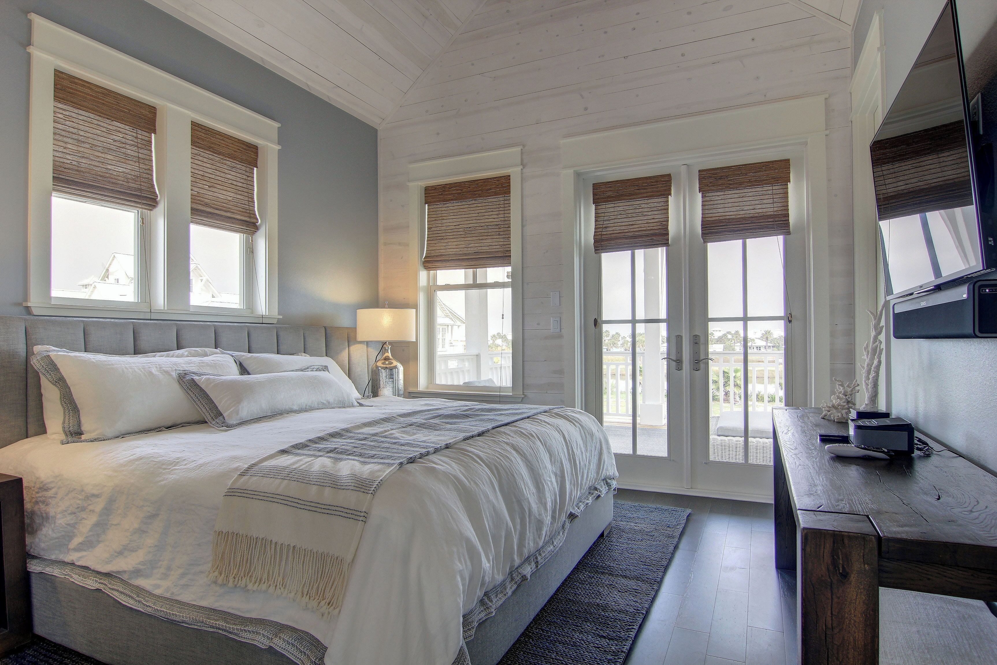 A bedroom with a large bed, a desk, and a TV. There are windows with blinds and a view of the outside.
