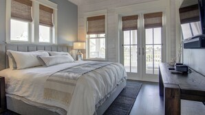 A bedroom with a large bed, a desk, and a TV. There are windows with blinds and a view of the outside. - Coastal Charm 258 HI (Port Aransas)