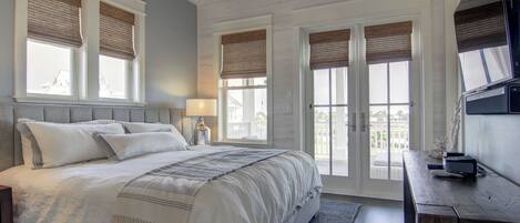 A bedroom with a large bed, a desk, and a TV. There are windows with blinds and a view of the outside.