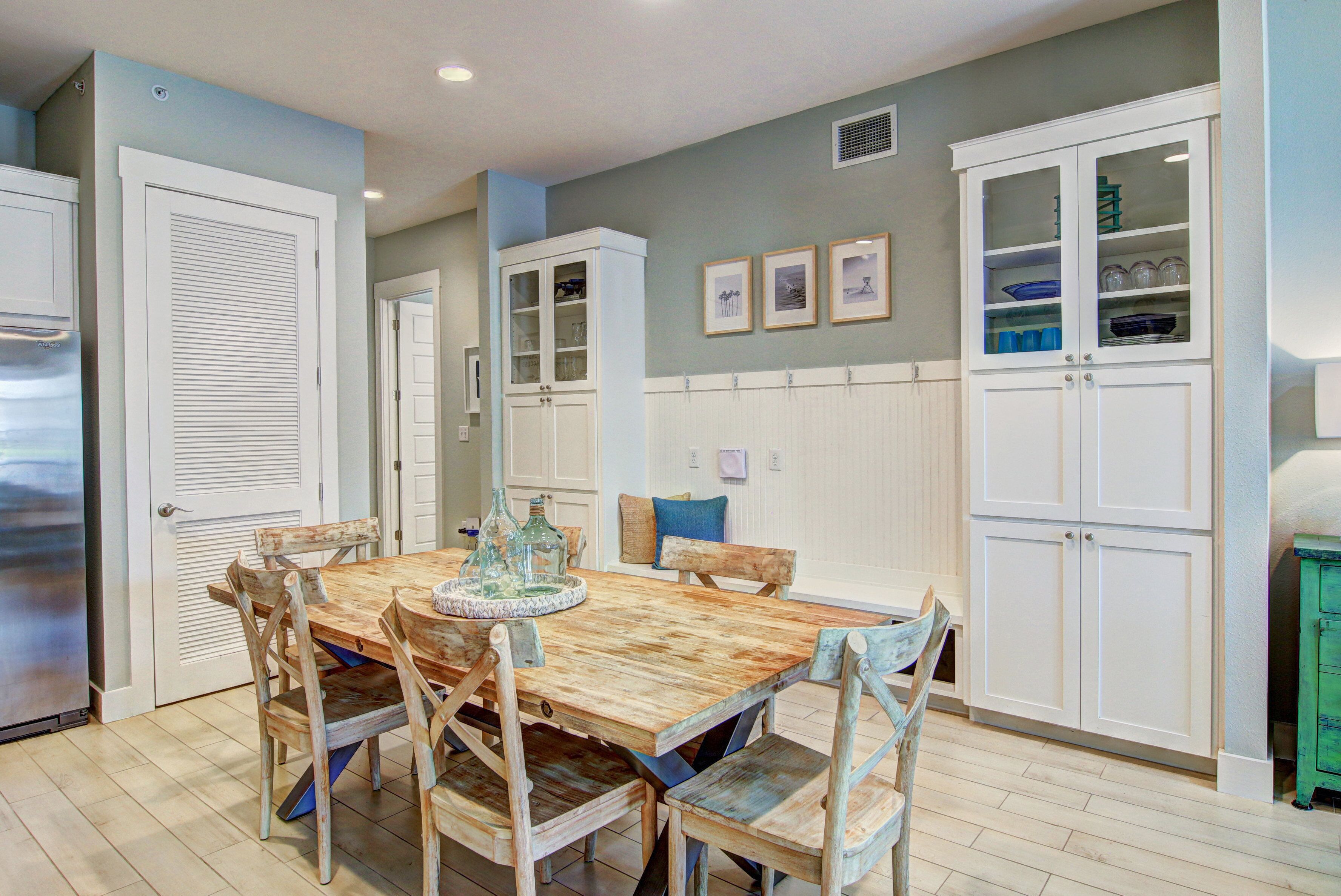 A dining area with a wooden table and chairs, white cabinetry, and a kitchen in the background.