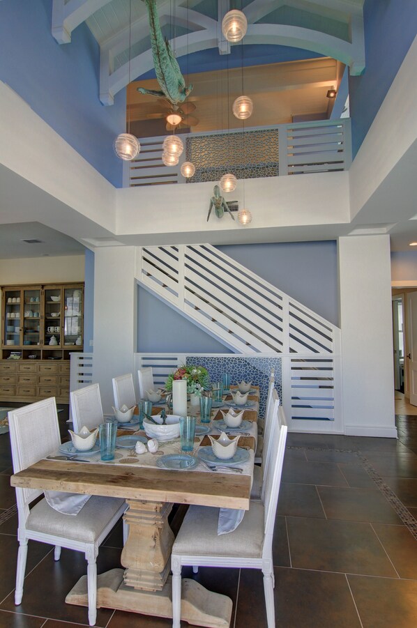 Enjoy meals in this dining area with a high ceiling and staircase, featuring a wooden dining table and comfortable seating.