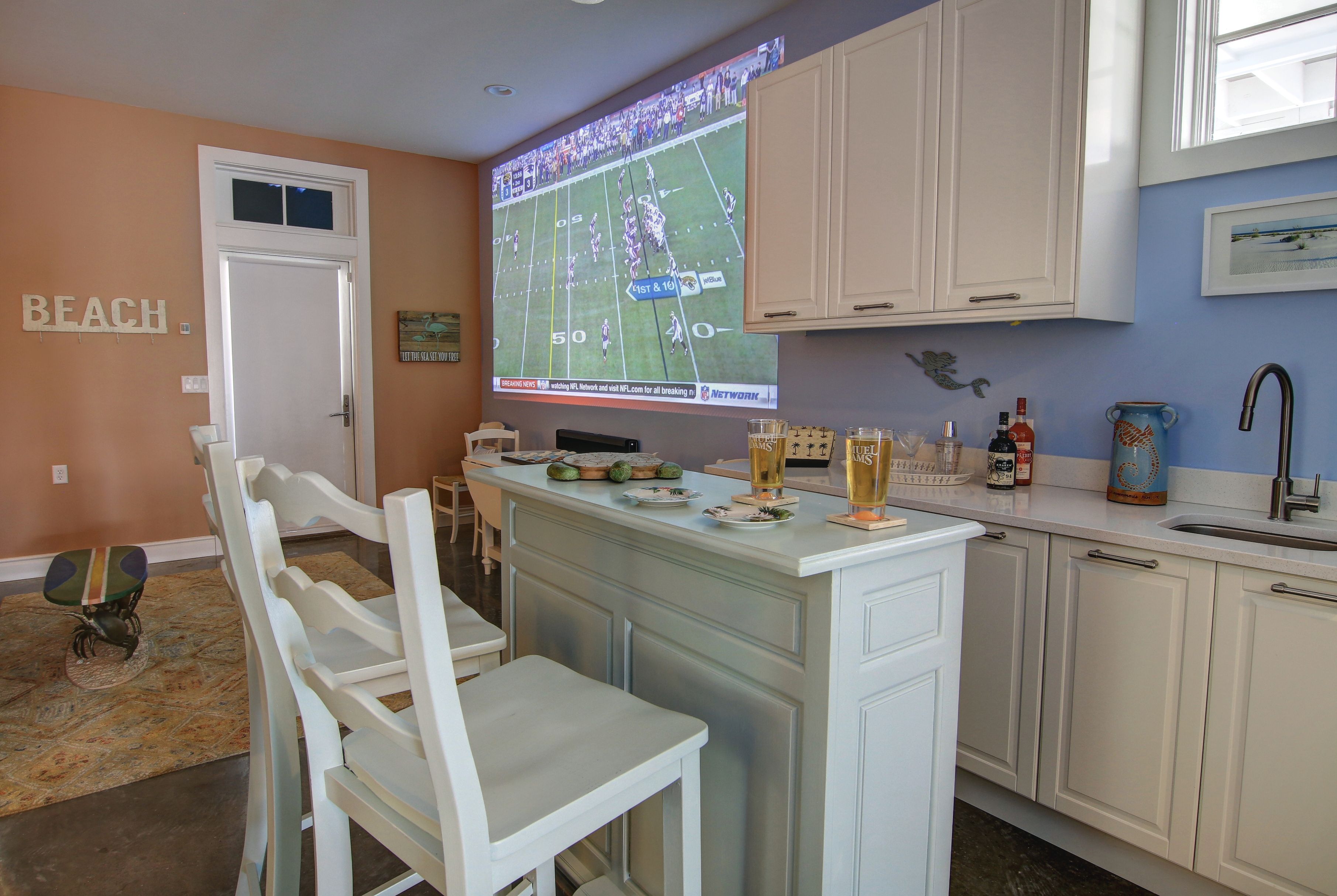 Una cocina con un televisor grande mostrando un partido de fútbol, una zona de bar con bebidas y una puerta que dice "BEACH".
