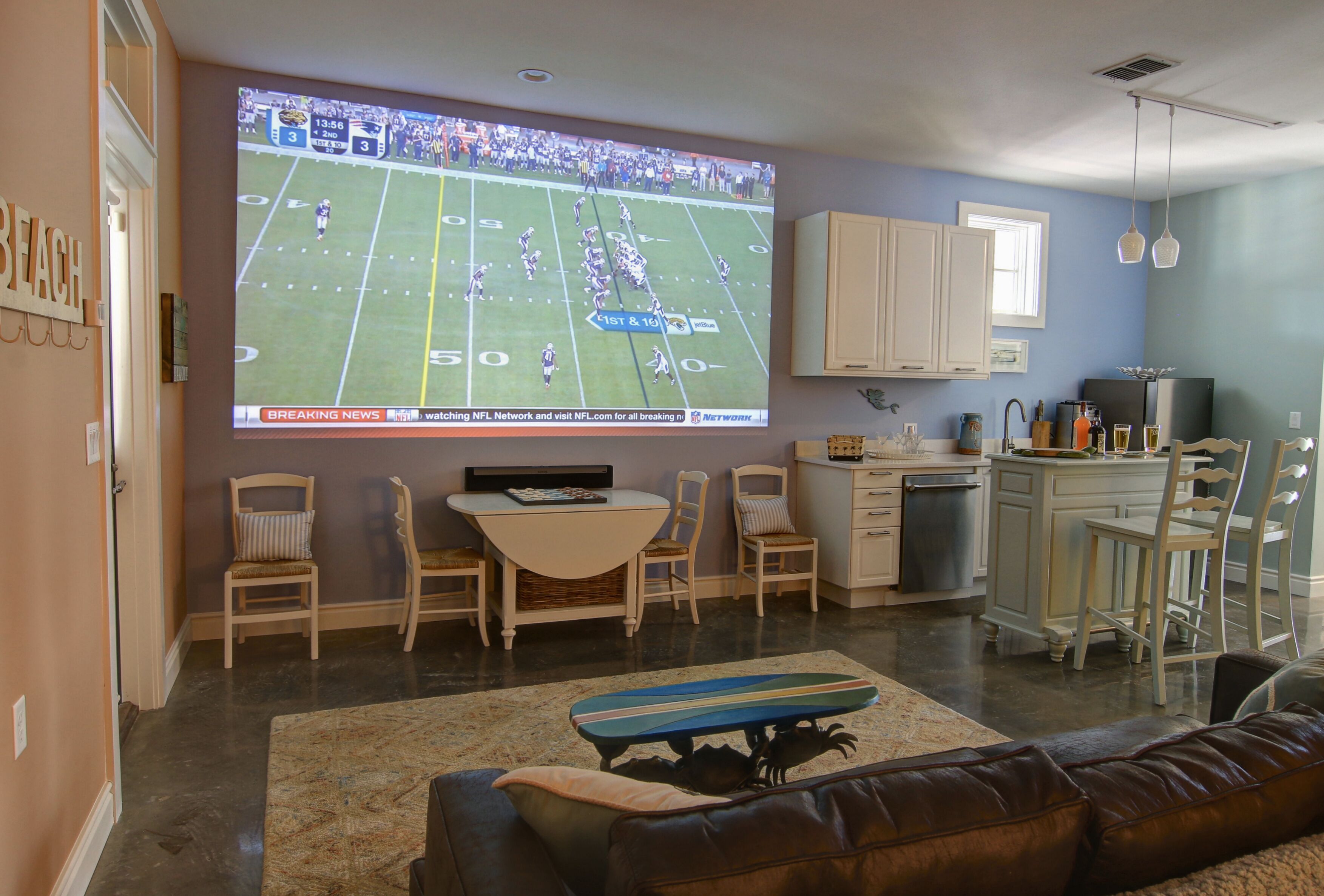 A living room with a large TV displaying a football game, a dining area with a table and chairs, and a kitchen area with cabinets and a refrigerator.