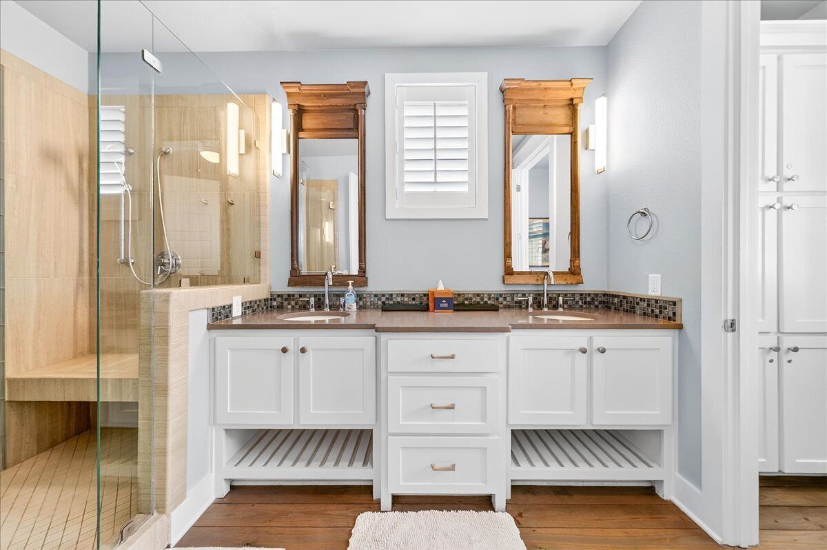 Une salle de bain dotée d’une douche à paroi de verre, de deux vasques avec des plans de travail en bois, de meubles blancs et d’un pare-feu en carreaux.