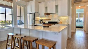 A kitchen with a large island, wooden stools, white cabinets, and a view through the window.