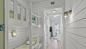 A narrow hallway with white wooden walls and a sloped ceiling. There are bunk beds with green and white patterned bedding, a small table, and a wall-mounted light fixture. - Nautical Launch TC 1207 (Port Aransas)