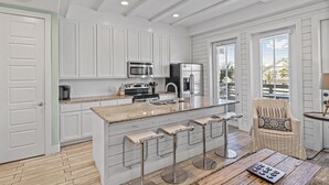 A modern kitchen with white cabinetry, stainless steel appliances, a central island with bar stools, and a view of the outdoors through a large window. - Nautical Launch TC 1207 (Port Aransas)