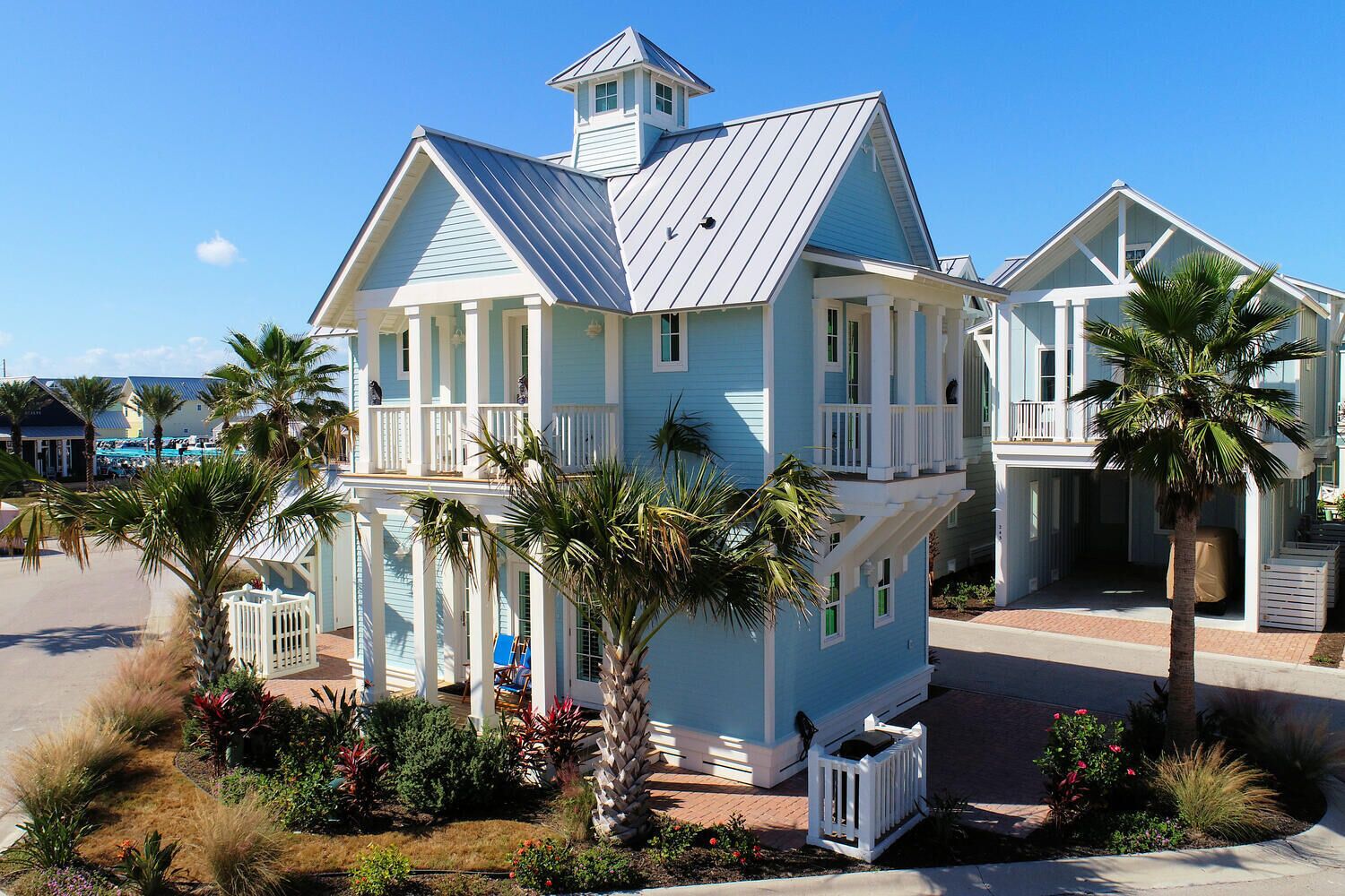 A large, light blue building with multiple balconies, surrounded by palm trees and a well-maintained garden.