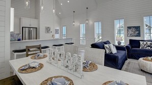 A spacious living area with a dining table set for four, a kitchen with white cabinetry, and a large window providing natural light. - Key Lime Cottage 248 BR (Port Aransas)