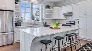 A modern kitchen with white cabinetry, a large island with stools, stainless steel appliances, and a window providing natural light. - One Particular Harbor 119 FI (Port Aransas)