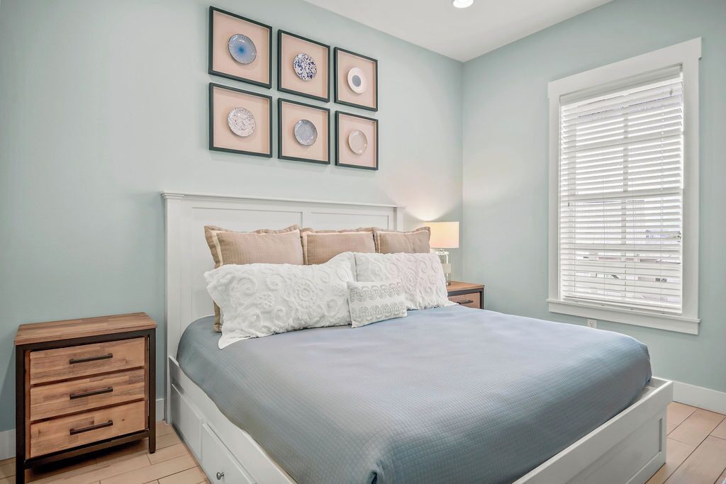 Une chambre à coucher comprenant un lit, des tables de chevet, une fenêtre avec des stores et des œuvres d’art encadrées accrochées au mur.