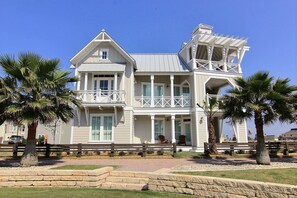 A two-story house with a balcony and a porch, surrounded by palm trees and a stone wall. - Oceanaire 196 RO (Port Aransas)