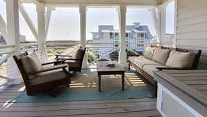 A covered outdoor seating area with a wooden table and two sofas, overlooking a large house and a beach. - Oceanaire 196 RO (Port Aransas)