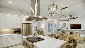 Una cocina moderna con armarios blancos, electrodomésticos de acero inoxidable, una isla central y una zona de comedor con mobiliario de mimbre.