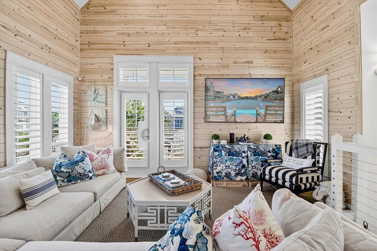 A cozy, wooden-paneled room with a sofa, armchairs, and a coffee table. There are two doors with shutters, a painting of a coastal scene, and a small table with a plant.