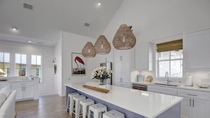 A modern kitchen with white cabinetry, a large island with bar stools, pendant lights, and a view of the outside through large windows. - Seas the Day 131 WI (Port Aransas)