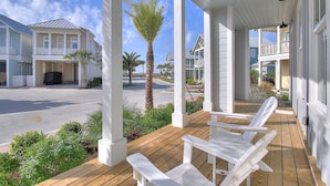 A covered porch with white chairs and a bench, overlooking a street with houses and palm trees. - Seas the Day 131 WI (Port Aransas)
