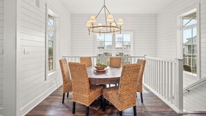 A dining area with a round table, wicker chairs, a chandelier, and a view through large windows. - Summer Catch 401 COA (Port Aransas)