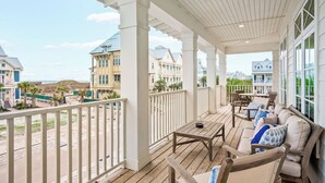 A covered outdoor area with wooden flooring, a seating area with cushions, and a view of beachfront buildings. - Summer Catch 401 COA (Port Aransas)