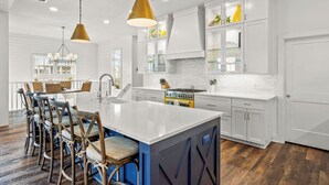 A modern kitchen with white cabinetry, a large island, and a dining area with wooden chairs. - Summer Catch 401 COA (Port Aransas)