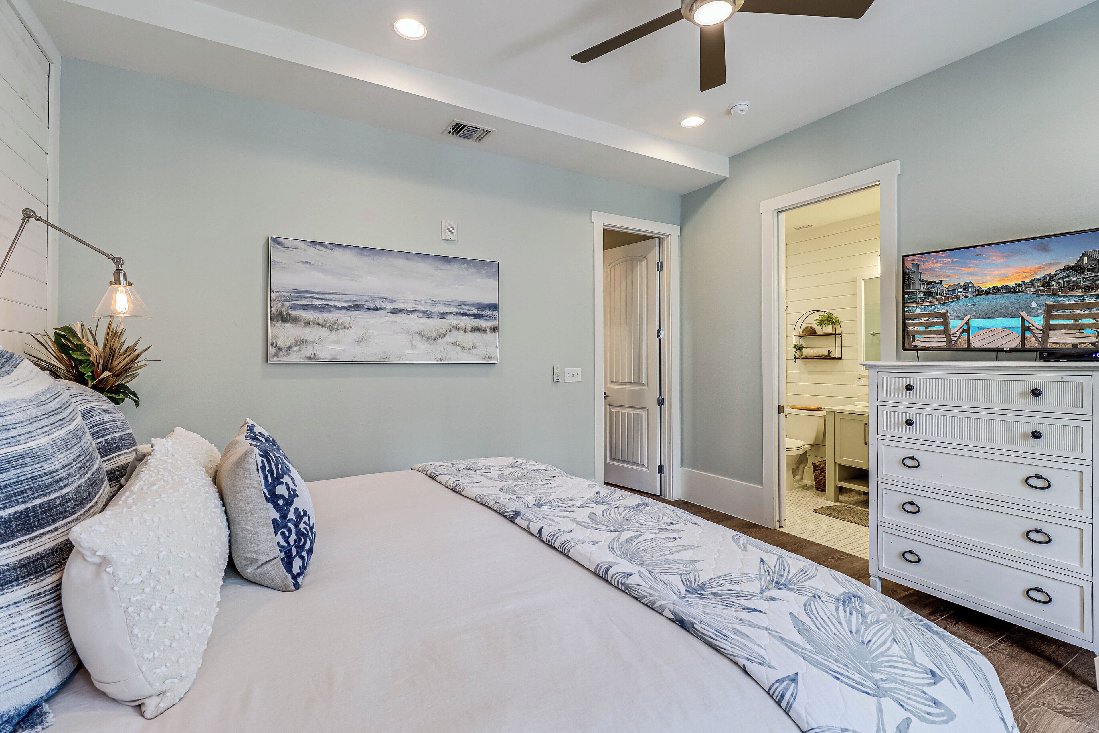 Un dormitorio con cama, cómoda, ventilador de techo, iluminación empotrada, un cuadro y una puerta que da a otra habitación.