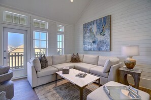 Una sala de estar con un sofá modular, una mesa de centro y una mesita de noche con una lámpara. Hay un ventanal de varios paneles y se ve una terraza al exterior.