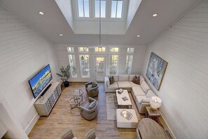 A spacious living room with a large TV, comfortable seating, and a view of the outdoors through large windows.