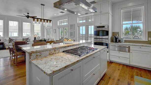 A modern kitchen with white cabinetry, a large island with a granite countertop, stainless steel appliances, and a view of palm trees through the window. - Sippin' on Sunshine 164 BE (Port Aransas)