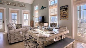 A dining area with a large table set for six, surrounded by four chairs. There is a television on a stand against the wall, and a decorative fish mounted above it. - Latitude Adjustment TC 3305 (Port Aransas)