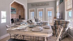 A dining area with a large table set for six, surrounded by upholstered chairs. There is a cozy seating area with a sofa and pillows in the background. - Latitude Adjustment TC 3305 (Port Aransas)