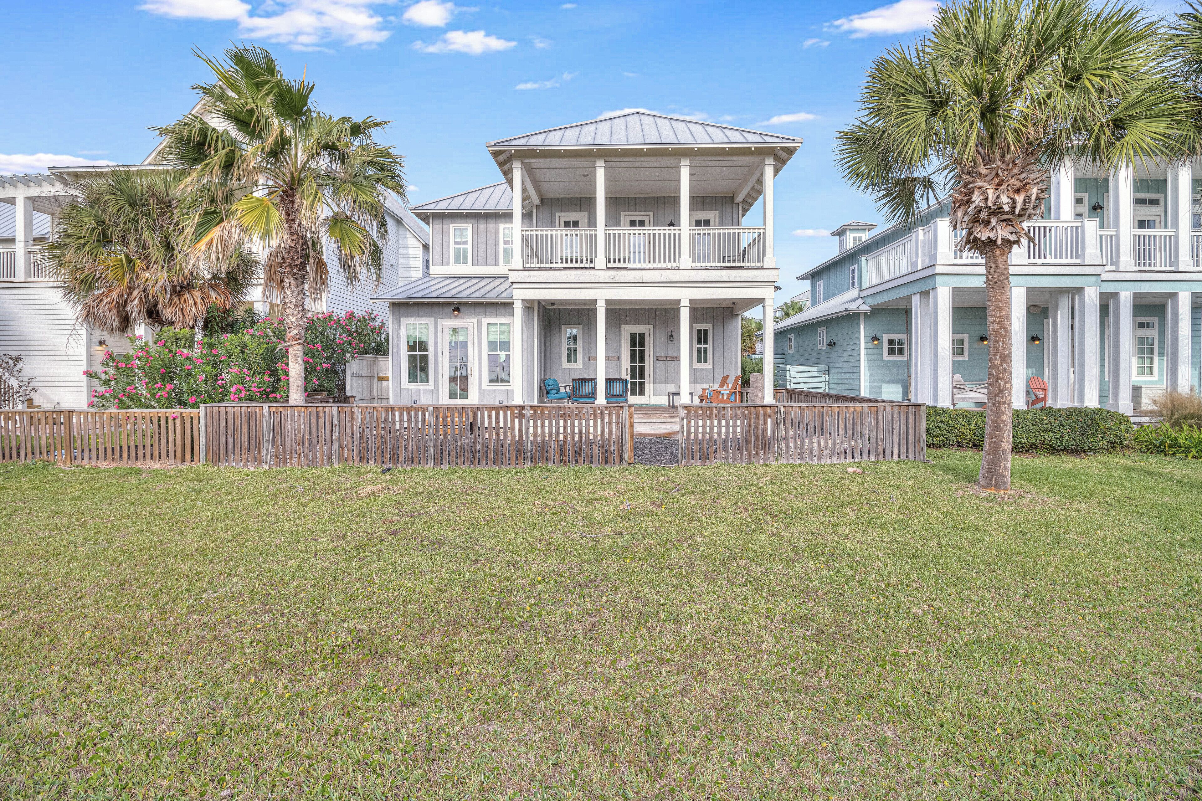 A two-story house with a balcony, surrounded by a wooden fence and palm trees.