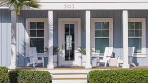 A two-story house with a balcony, numbered 303, featuring a front porch with chairs and a small garden area. - Sand In My Boots 303 HI (Port Aransas)
