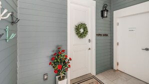 A hallway with a white door, a green wreath, a red flower arrangement, a wall-mounted lantern, and a doormat. - The Hideout TC 10101 (Port Aransas)