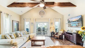 Un salon spacieux avec un plafond haut, un ventilateur de plafond, une grande fenêtre, un canapé, une table basse et un téléviseur.