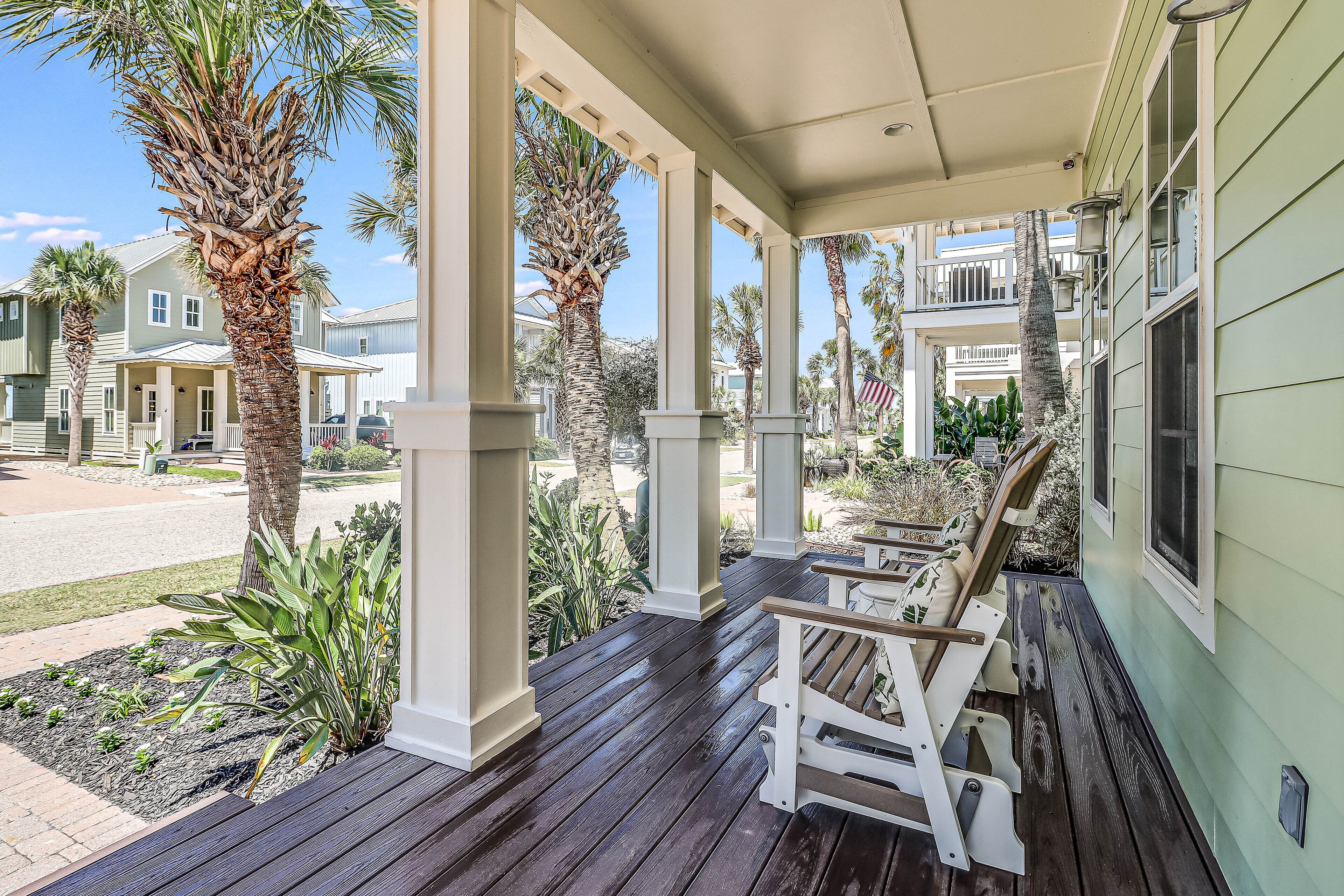 A covered porch with wooden benches, palm trees, and a view of neighboring houses.