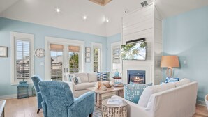 A living room with a white sofa, blue armchair, and a fireplace. There are windows with blinds, a clock, and a television on the wall. - Shore House 183 BE (Port Aransas)