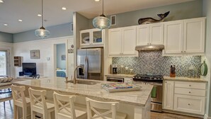 A modern kitchen with white cabinetry, a central island with bar stools, a stainless steel refrigerator, and a tiled backsplash. - Watercolor Bungalow TC 2101 (Port Aransas)