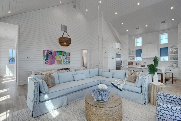A spacious living room with a light blue sectional sofa, a round coffee table, and a kitchen area in the background. - See The Sea 116 TUC (Port Aransas)