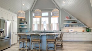 A kitchen with white cabinets, a central island with bar stools, pendant lighting, and a view of the outdoors through large windows. - The Salty Dog 314 GR (Port Aransas)