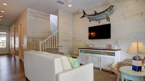 A living room with a white sofa, a dining area with a table and chairs, a mounted fish on the wall, and a staircase leading to another level. - Suits Us 231 BE (Port Aransas)