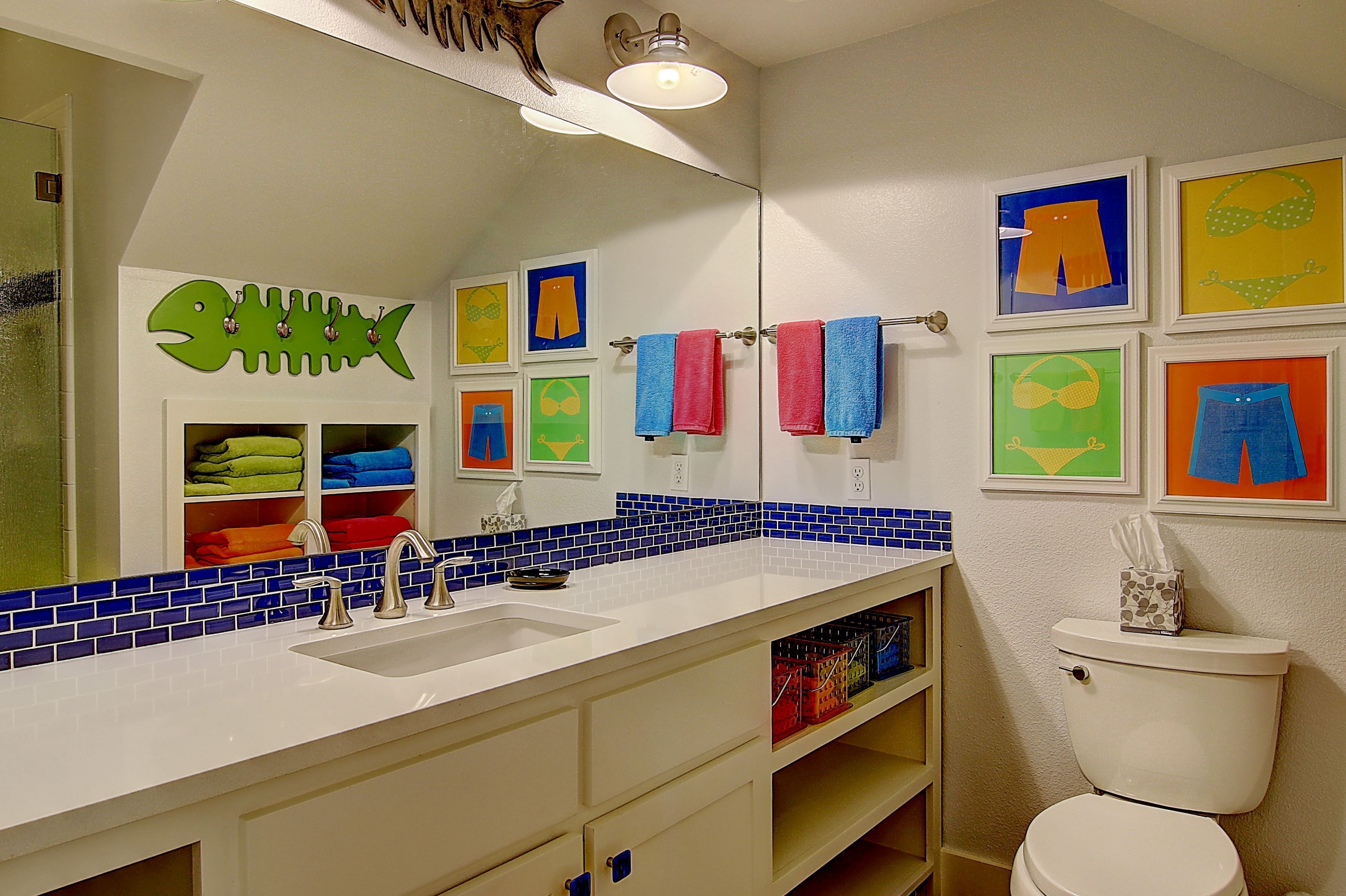 Une salle de bain avec un lavabo blanc, un mur recouvert de carreaux bleus derrière le lavabo et des toilettes. Des serviettes colorées et des œuvres d’art ornent les murs.