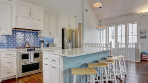 A kitchen with white cabinets, a stainless steel refrigerator, a double oven, and a blue backsplash. There are wooden stools at the island and a chandelier hanging from the ceiling. - Suits Us 231 BE (Port Aransas)