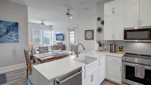 A modern kitchen with white cabinetry, stainless steel appliances, and a dining area with a large window. - Seaside TC 10301 (Port Aransas)