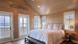 Un dormitorio con techo de madera, una cama grande, un banco y vistas a través de las ventanas.