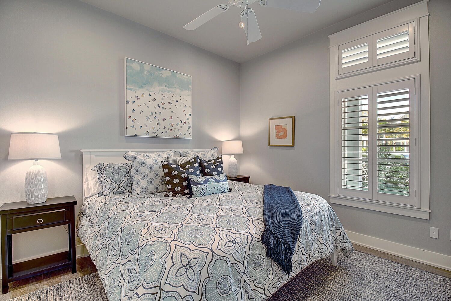 Une chambre à coucher avec une couvre-lit à motifs, deux lampes de chevet, un ventilateur de plafond et une grande fenêtre avec des stores.