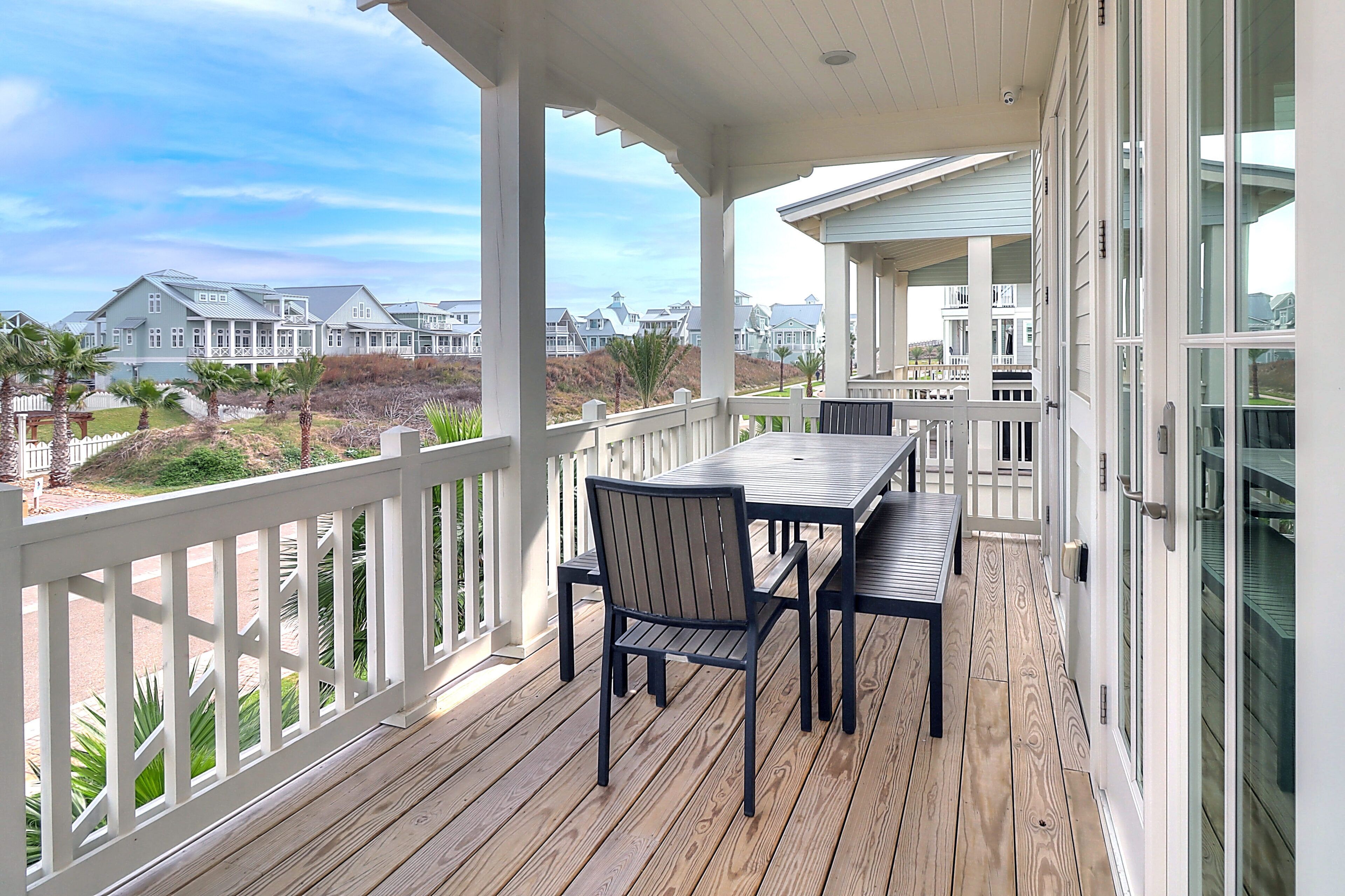 Un espace extérieur abrité, équipé d’une table et de chaises, qui offre une vue sur un quartier résidentiel avec ses maisons et une plage.