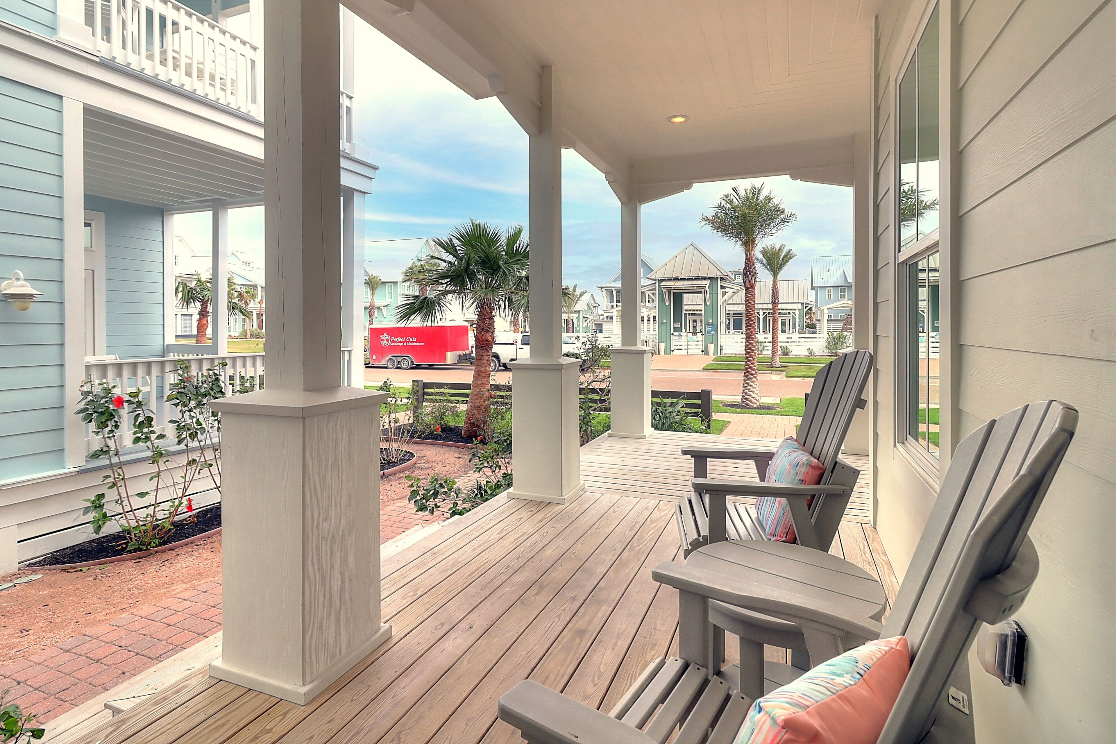 A covered porch with wooden flooring, a seating area with chairs and cushions, and a view of the street with palm trees and a red delivery truck.